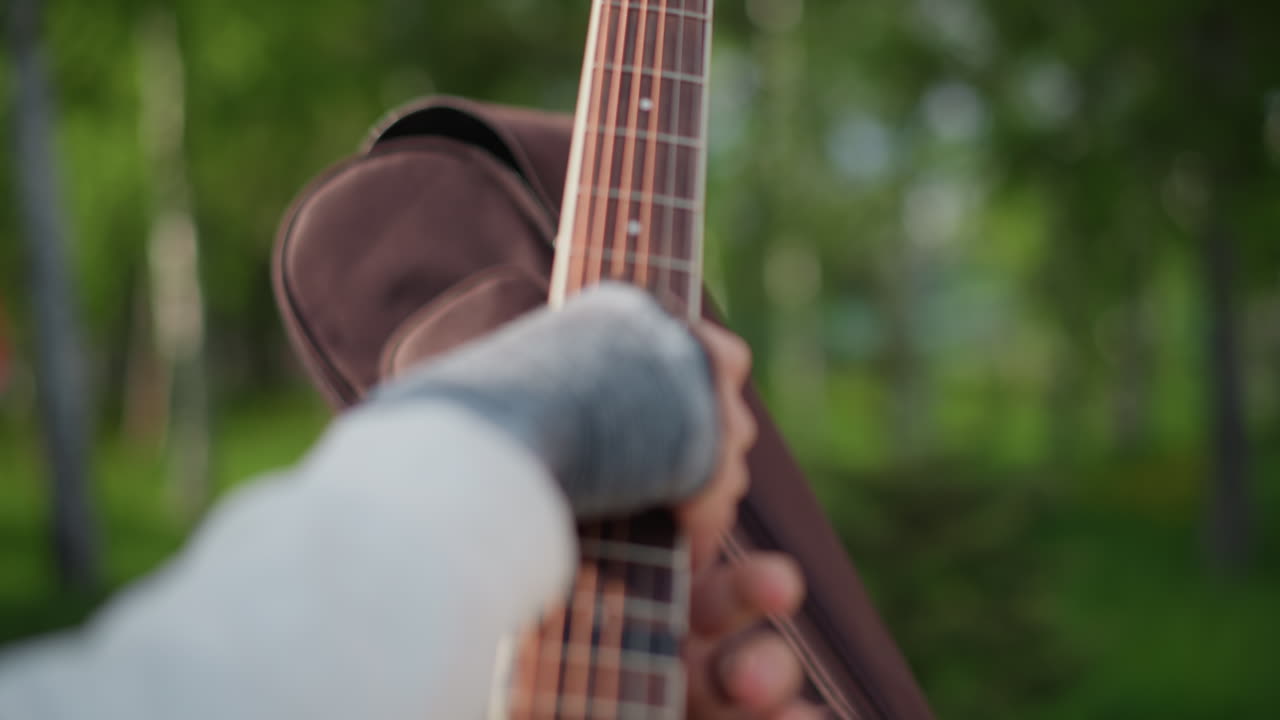 Dłoń zamyka futerał gitary na ławce, widoczna rękawica bez palców i dżinsowy rękaw, zamek błyskawiczny zabezpiecza wyściełaną torbę, muzyk wsuwa gitarę do futerału, w tle brzozy, ciepłe światło dzienne, moment