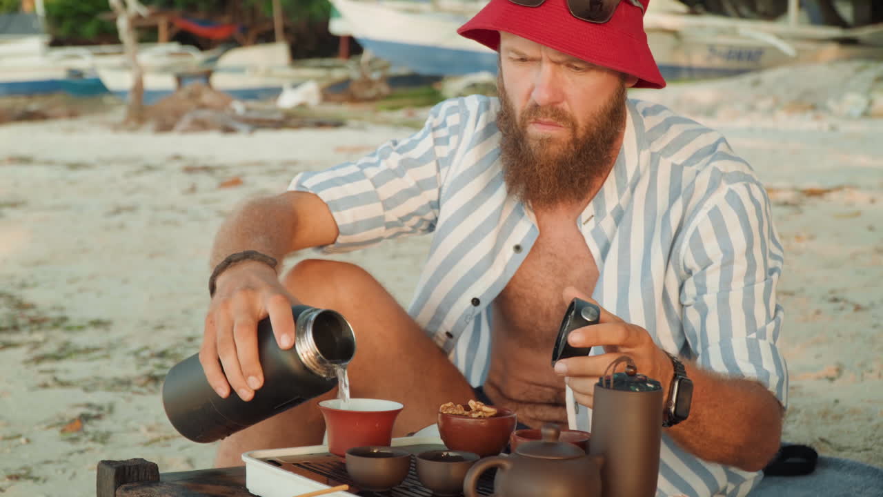 Cerimonia del tè in spiaggia con viaggiatore barbuto che versa il tè da un thermos in piccole tazze di ceramica sulla riva sabbiosa, uomo con cappello da pescatore rosso e camicia a righe che sistema teiera e vassoio accanto a legni trasportati dal mare e ancorato
