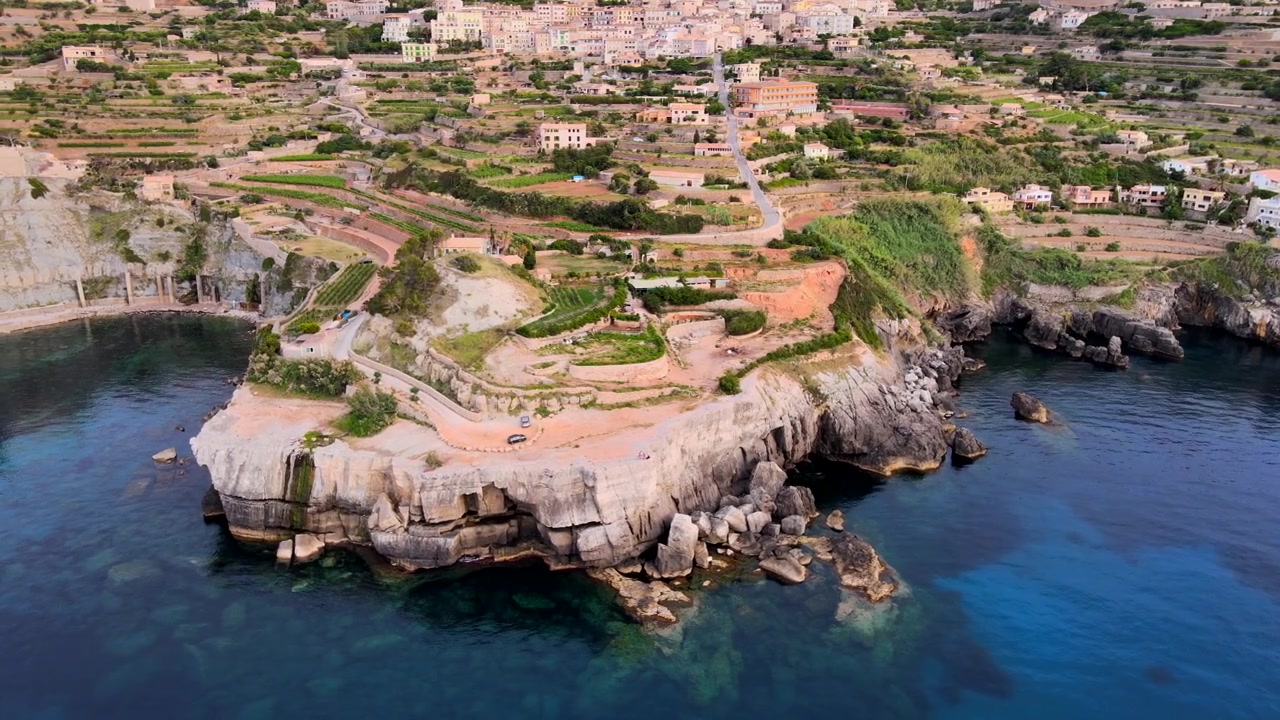 En betagende panoramaudsigt over en lille by på klipperne på Mallorca, omgivet af frodige grønne terrasser. Middelhavet glitrer nedenfor og skaber en fantastisk kontrast til det barske landskab.