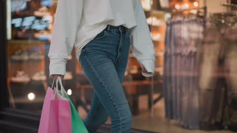 lower angle view of lady in jeans holding pink and mint shopping bags while walking past clothing store with blurred background featuring shoes, male and female clothes on racks