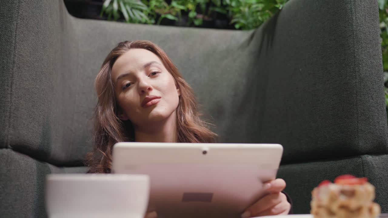 Lady seated in cozy green booth holds tablet and looks directly at camera with confident expression, while waffle and latte rest on wooden table, blending modern lifestyle with relaxed ambiance