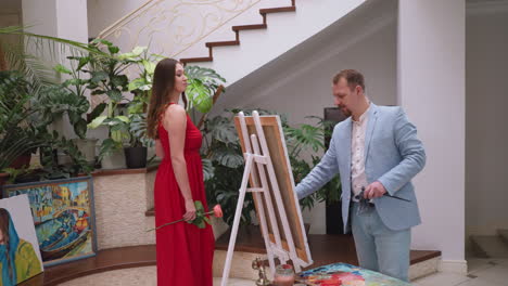 man draws standing live model on canvas. young woman in red dress poses for professional painter slowly approaching easel in art studio. artistic expression