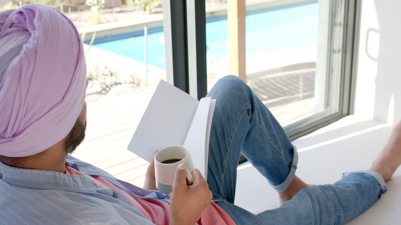 Relaxing at home, man in turban reading book and drinking coffee by window