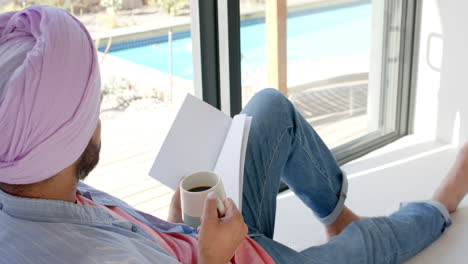 relaxing at home, man in turban reading book and drinking coffee by window