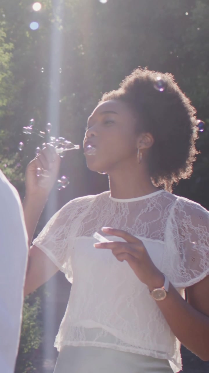 Vidéo verticale : Femme soufflant des bulles, portant une baguette à ses lèvres dans un parc pour s'amuser, tenant la solution et observant sa blouse.