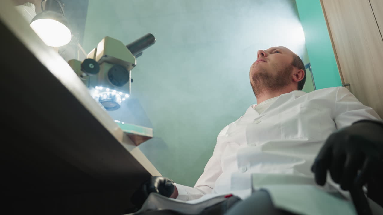 una vista hacia abajo de un técnico en una bata de laboratorio blanca, sentado y mirando pensativo en un laboratorio bien iluminado con la luz que brilla de la lámpara de escritorio y el microscopio y en la pared