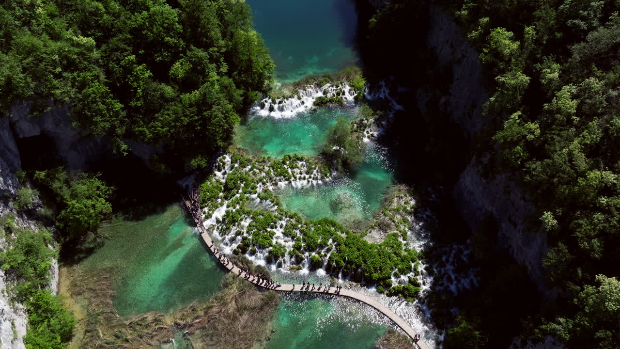 Tourists Walking On Path Through Turquoise Lakes At Plitvice Lakes National Park In Lika-Senj County, Croatia. aerial pullback shot