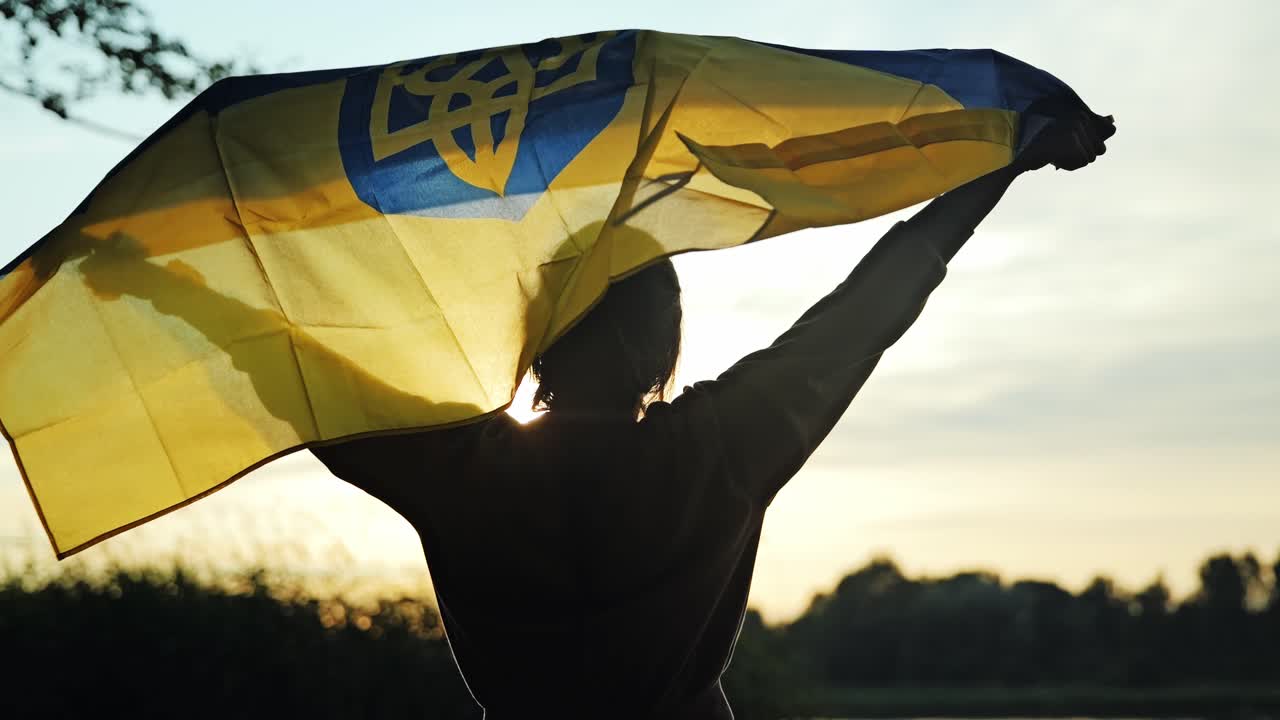 Woman lifts Ukrainian flag slowly, riverside sunset, embodying silent resilience