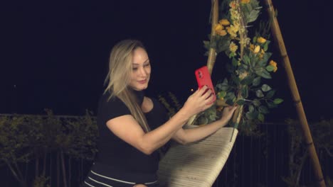 Hispanic-woman-posing-on-a-swing-chair-outside-at-nighttime-for-a-smartphone-selfie