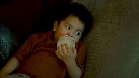 British-Asian-18-month-old-baby-boy-sits-leisurely-on-a-home-sofa,-holding-a-bottle-of-milk-and-drinking-from-it,-bathed-in-the-warm-glow-of-TV-screen-light-reflections