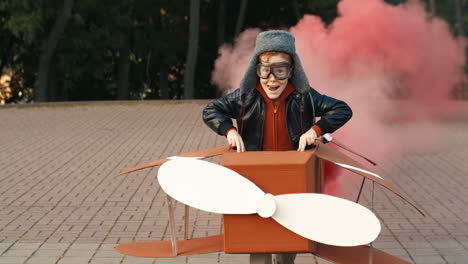 Cute-Little-Red-Haired-Boy-In-Hat-And-Glasses-Running-In-The-Park-Holding-A-Cardboard-Plane-With-Smoke-Behind-And-Playing-As-An-Aviator