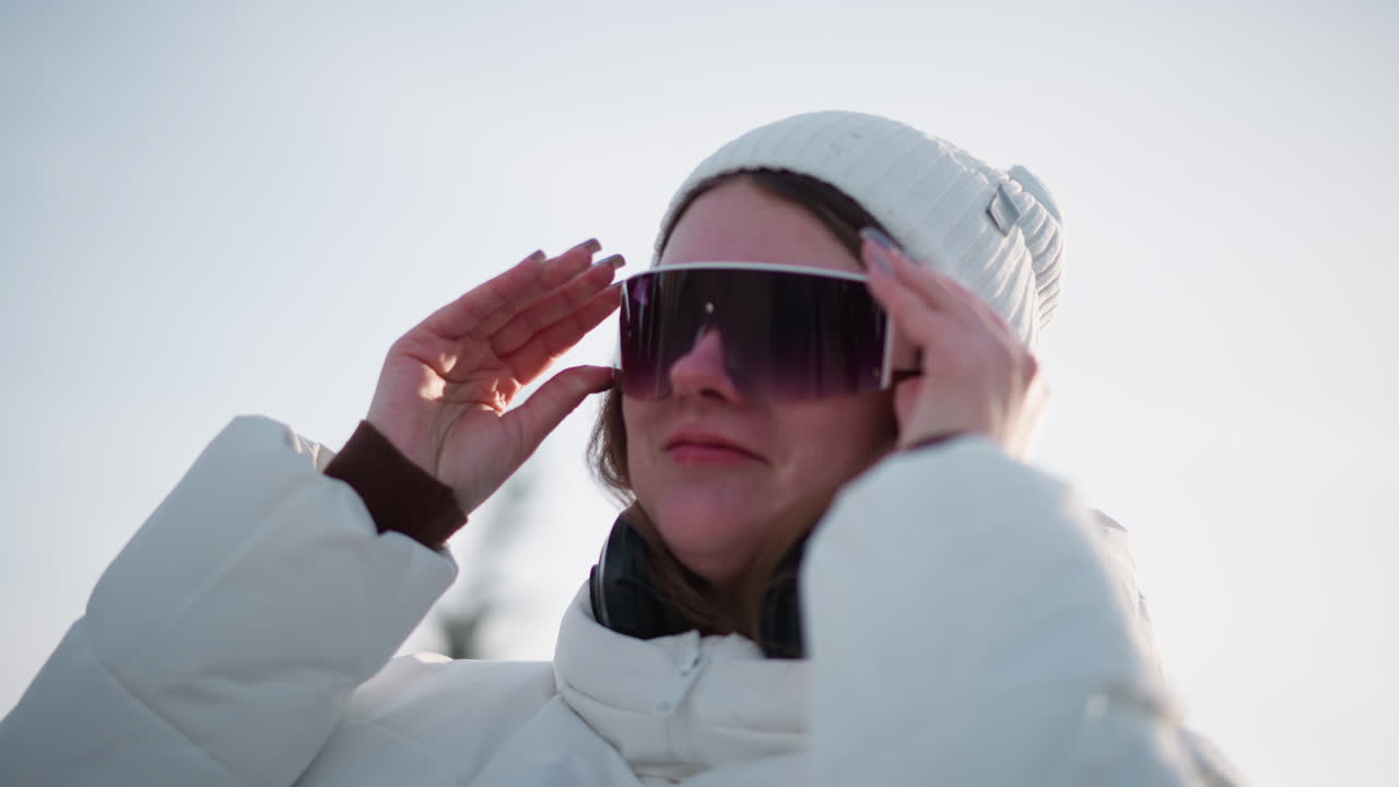 Close up of radiant lady with rosy cheeks donning sunglasses under bright sky while smiling joyfully, breeze tousles hair, light glints on lenses, headphones on neck, wearing coat and beanie