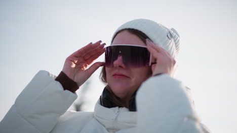 close up of radiant lady with rosy cheeks donning sunglasses under bright sky while smiling joyfully, breeze tousles hair, light glints on lenses, headphones on neck, wearing coat and beanie