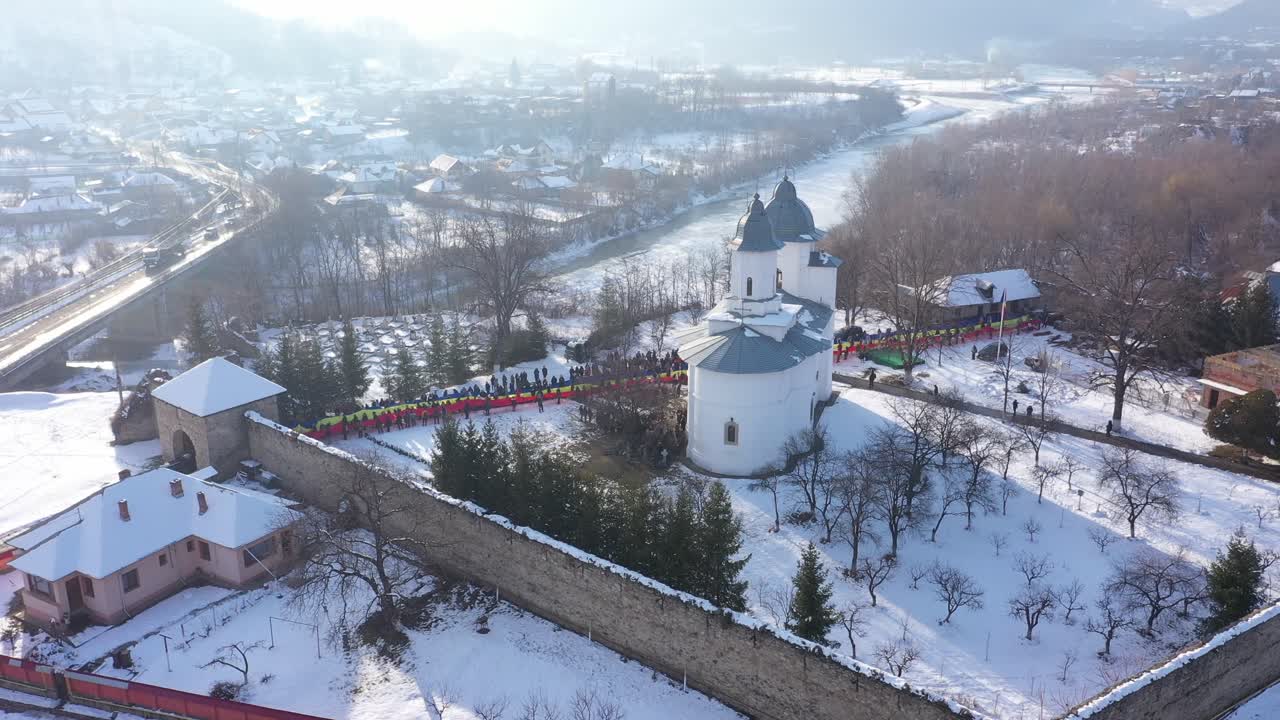 An ascending aerial shot slowly reveals Raducanu Monastery in Targu Ocna, Romania. A long line of people holds a massive Romanian flag on the snow-covered grounds during a winter national event