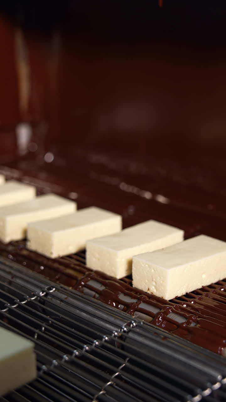 White candies move on the conveyor line to the flow of chocolate. Manufacture of chocolate candies at the sweets factory. Close up. Vertical video