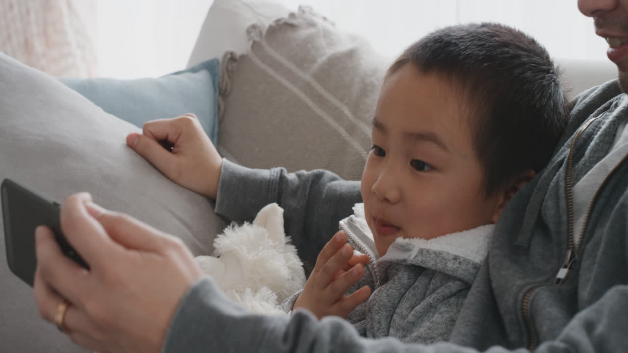 행복한 아시아 소년과 아버지가 스마트폰을 사용하여 휴대폰 터치 스크린 장치에서 온라인 엔터테인먼트를 시청하고 인터넷을 검색하고 소파에서 함께 휴식을 취합니다 4k 영상