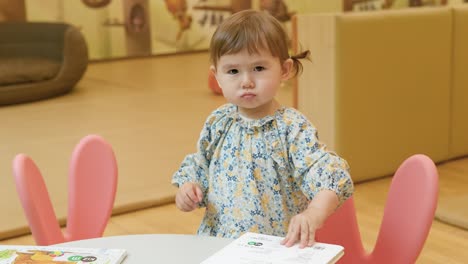 Adorable-Niña-De-2-Años-Se-Sienta-A-La-Mesa-Y-Mira-Un-Libro-Colorido-Con-Fotos-Y-Un-Libro-De-Cierre,-Se-Levanta-Y-Mira-La-Cámara-Con-Cara-Sabia