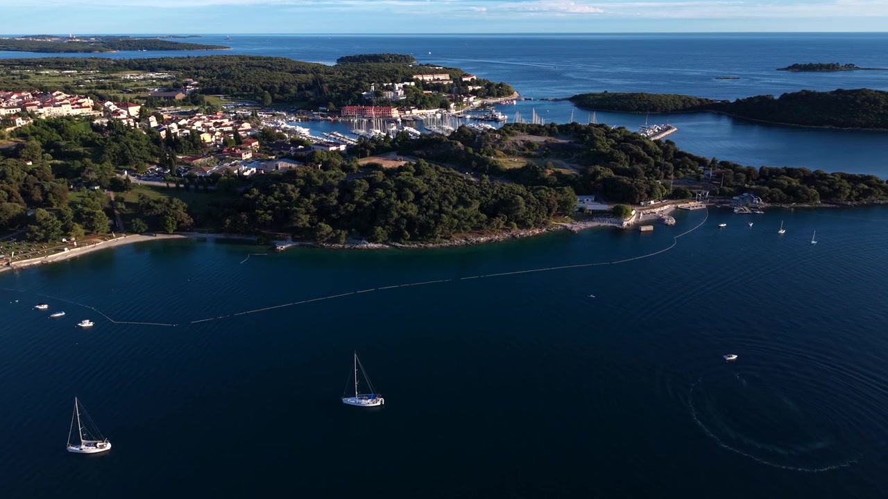 Barcos à vela flutuam pelo calmo Mar Adriático perto da histórica Vrsar, Croácia, Europa, emoldurado por ilhas arborizadas, cidade costeira, porto, iates à vela sob a luz dourada do pôr do sol. Tomada aérea de drone em aproximação.