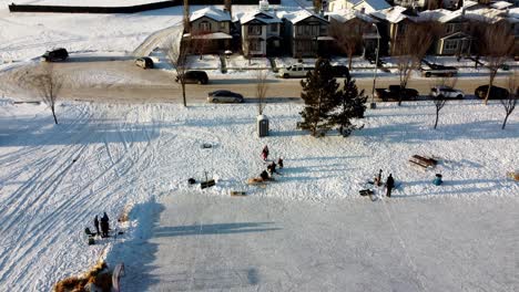 Elevación-Aérea-De-Invierno-Sobre-Comunidad-Residencial-Pista-De-Hielo-Hecha-Por-El-Hombre-Gente-Practicando-Patinaje-Hockey-Y-Aprendiendo-A-Patinar-Parque-Comunitario-Fuera-De-Casa-Gente-Comunicándose-En-Mesa-De-Picnic-Pozo-De-Fuego-Montones-De-Heno-1-3