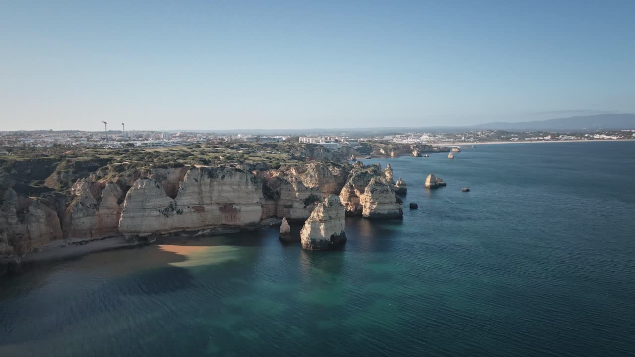 Camera spirals along sunlit cliffs and turquoise waters, revealing the intricate coastline, rugged beaches, and distant Portuguese town