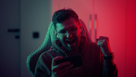 a young man with glasses a professional gamer on a mobile phone plays games and emotionally rejoices in the victory. emotional young man playing mobile games in a neon light. high quality 4k footage