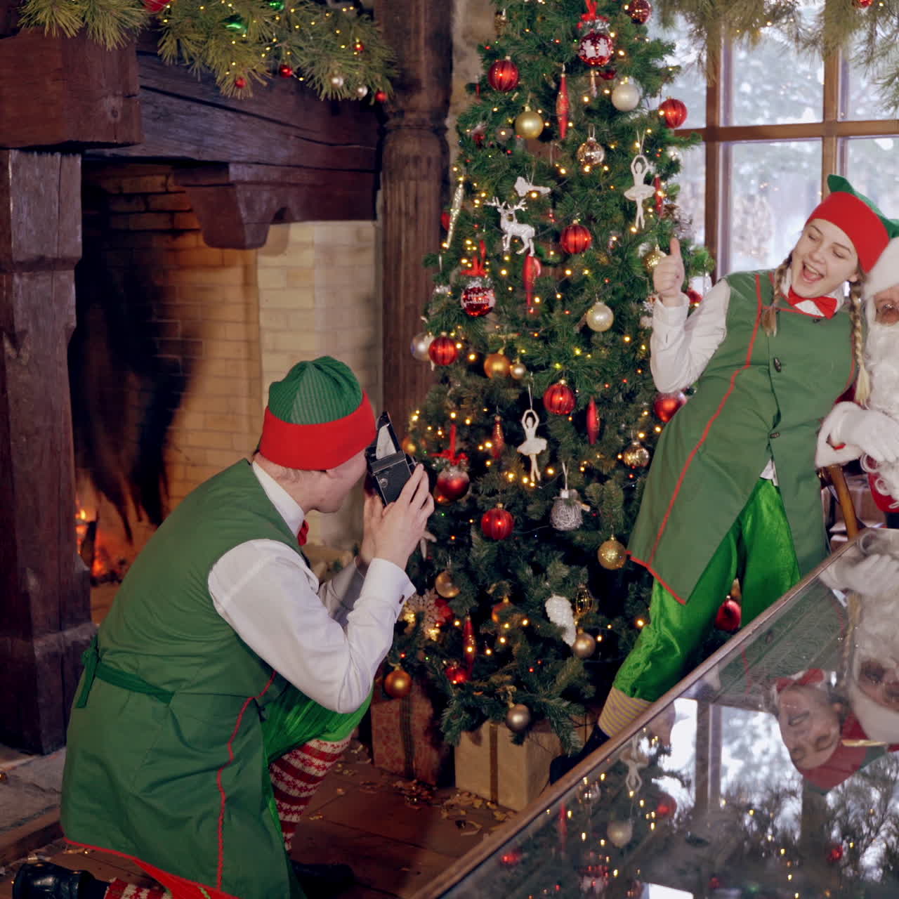 Elf boy is taking picture of Santa Claus and girl elf near the NewYear tree in decorated room. Capturing moments of happiness