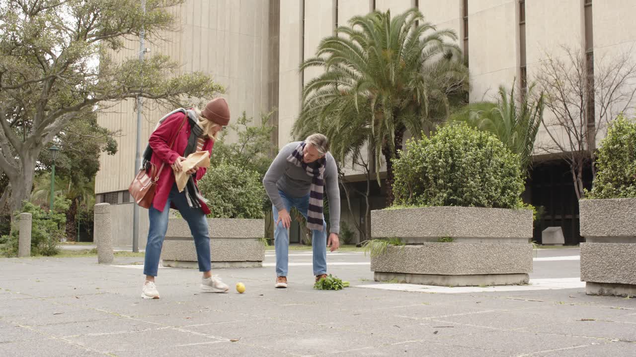 Coppia anziana che cammina in piazza, si ferma mentre la spesa cade dalla borsa, raccogliendo i prodotti.
