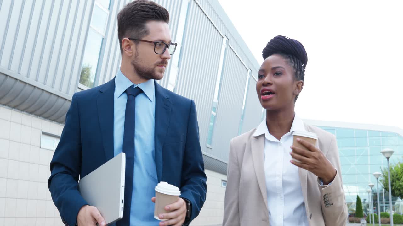 African american businesswoman and caucasian businessman holding coffee cups and talking while walking down the street