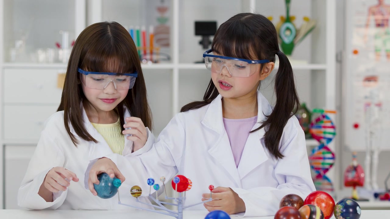 Two young girls wearing safety goggles and lab coats examine a planetary model in classroom.