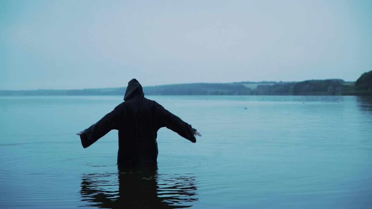 Evil witch in black cloak with hood in the river. Divination by the water. Halloween costume.