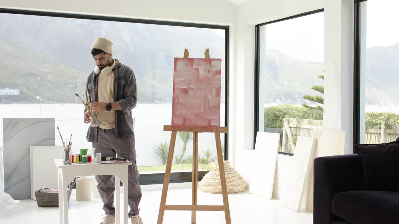 Abstrakt lærred, der inspirerer indisk mand i atelier ved søen, som inspicerer pensler og forbereder sig på at tegne