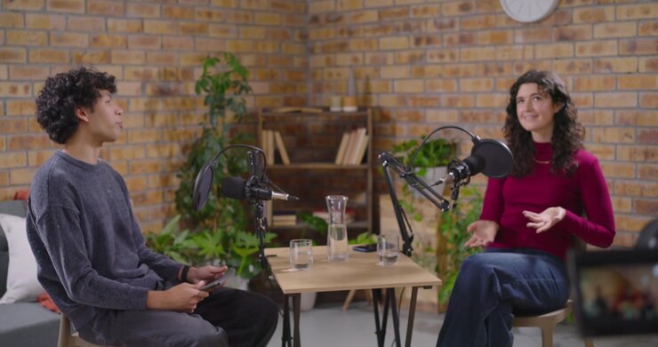 Two hosts speaking, gesturing at mics on set, camera operator adjusting framing to record interview