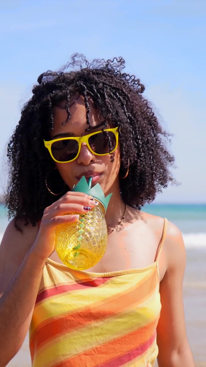 Happy Young Woman with Curly Hair Drinking from Pineapple Cup on Beach