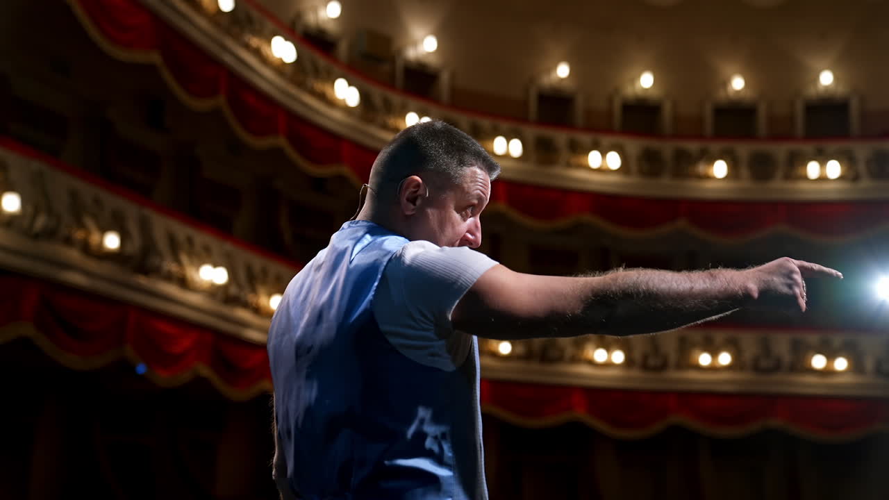 Presenter in front of empty theatre hall with red velvet chairs. Actor is standing on stage and talking with gestures before the performance.
