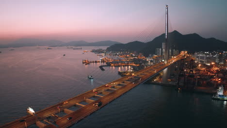 multiple cars and busses driving over the busy stonecutters bridge after sunset with oil depot and mountain range in the back drop