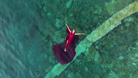 Vista-Aérea-De-Una-Mujer-Con-Un-Largo-Vestido-Rojo-Flotando-Sobre-Aguas-Turquesas-Del-Mar-Adriático