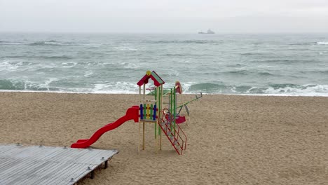 Einsamer-Verlassener-Bunter-Kinderspielplatz-Am-Leeren-Meeresstrand,-Schiff,-Das-Sich-Im-Hintergrund-Bewegt,-Einspielung