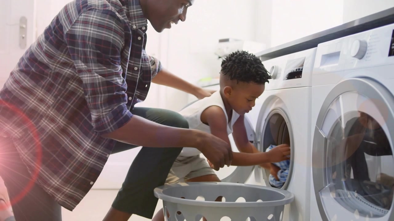 Father guiding son after child opening washer door, both loading garments for household laundry