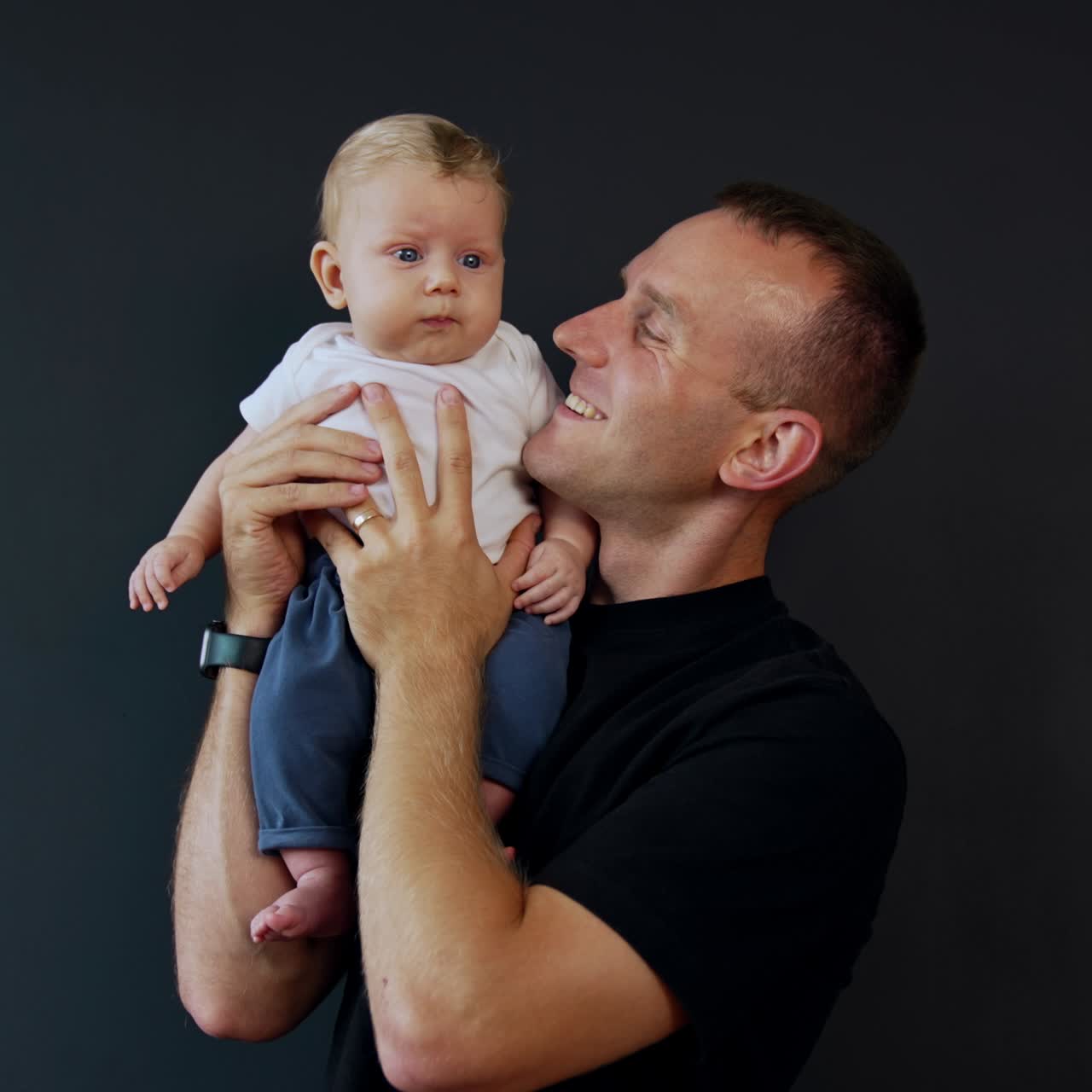 Uomo felice con una t-shirt nera che tiene il suo neonato sulla spalla. Il bambino si guarda intorno e il padre lo bacia sulla guancia.