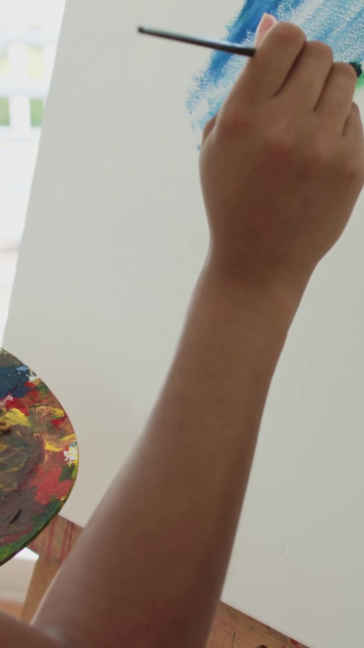 Vídeo vertical: Pintor(a) mergulhando pincel na paleta, misturando azul-petróleo e adicionando camada azul em tela de pintura em casa