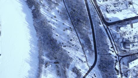 Vista-Aérea-De-Pájaros-Sobre-El-Tráfico-Ligero-De-La-Ciudad-De-Invierno-Por-Un-Río-Cubierto-De-Nieve-Congelada-Al-Lado-De-La-Carretera-Por-Una-Zona-Residencial-De-La-Comunidad-De-La-Ciudad-Con-Una-Carretera-Que-Conduce-A-La-Zona-Forestal-De-La-Autopista