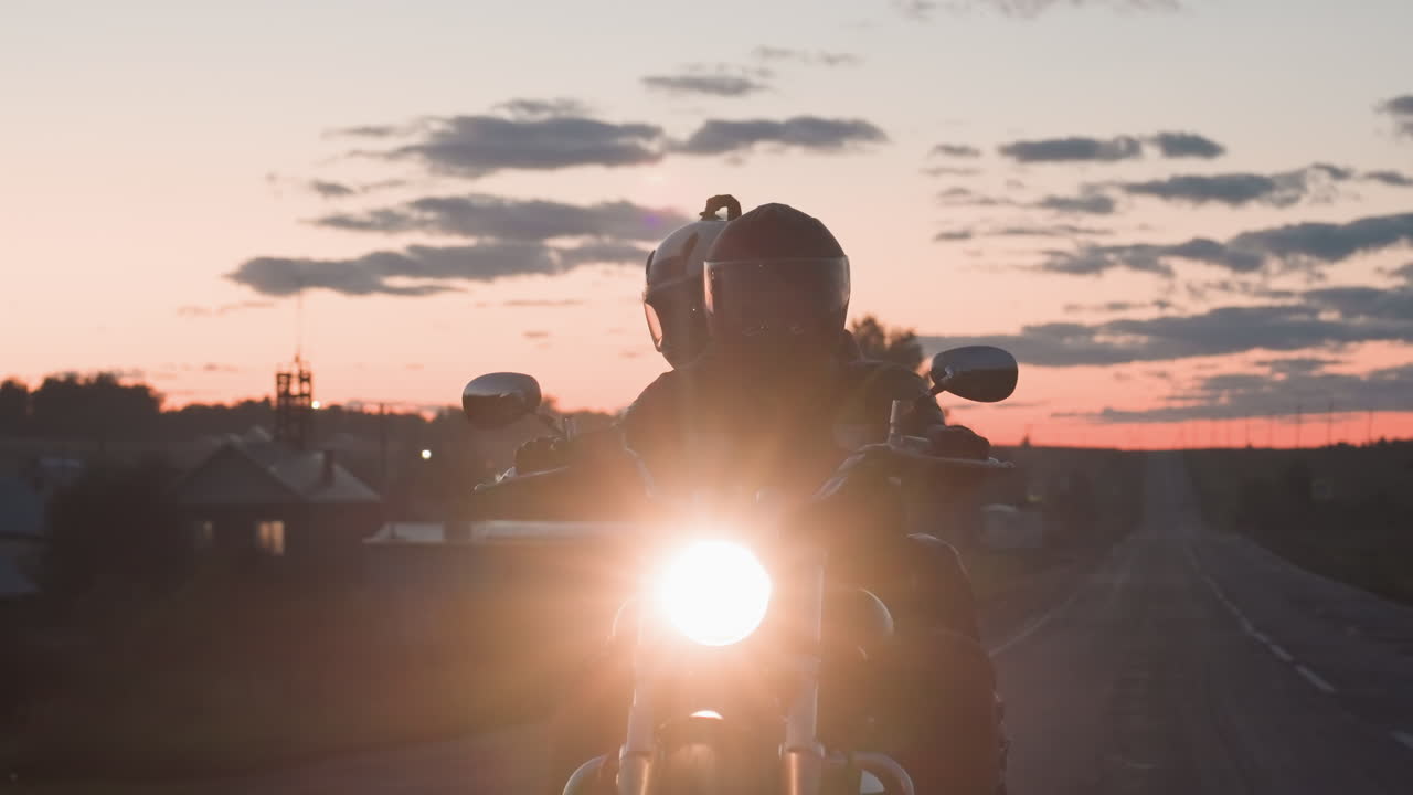 Night rider carrying passenger cruising countryside road under glowing headlamp with helmets on, evening sky filled with dramatic clouds and fading sunset casting warm light across landscape