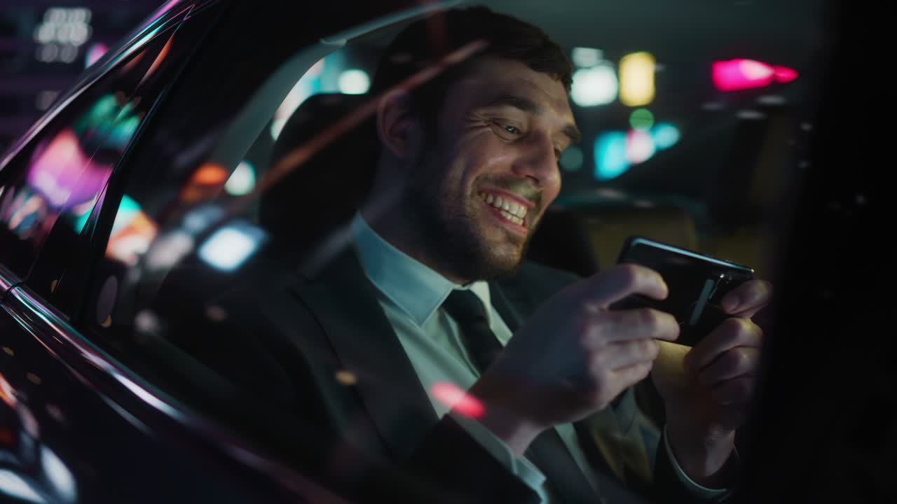 Businessman in a Suit Commuting from Office in a Backseat of His Car at Night. Entrepreneur Playing Video Games on Smartphone and Winning. Urban City Street with Working Neon Signs.