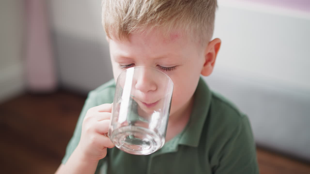 Close up of kid in green top drinking water while holding clear glass cup with both hands, sitting in naturally lit indoor space, expressing calmness