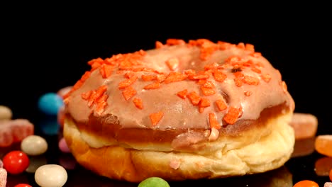 orange funny surprised glazed donut with sprinkles, striped caramel candies, colorful dragee with raisins or peanuts inside rotate on black background