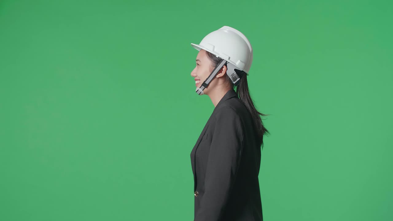 Side View Of Smiling Asian Female Engineer Looking Around And Shaking Her Head While Walking In The Green Screen Studio