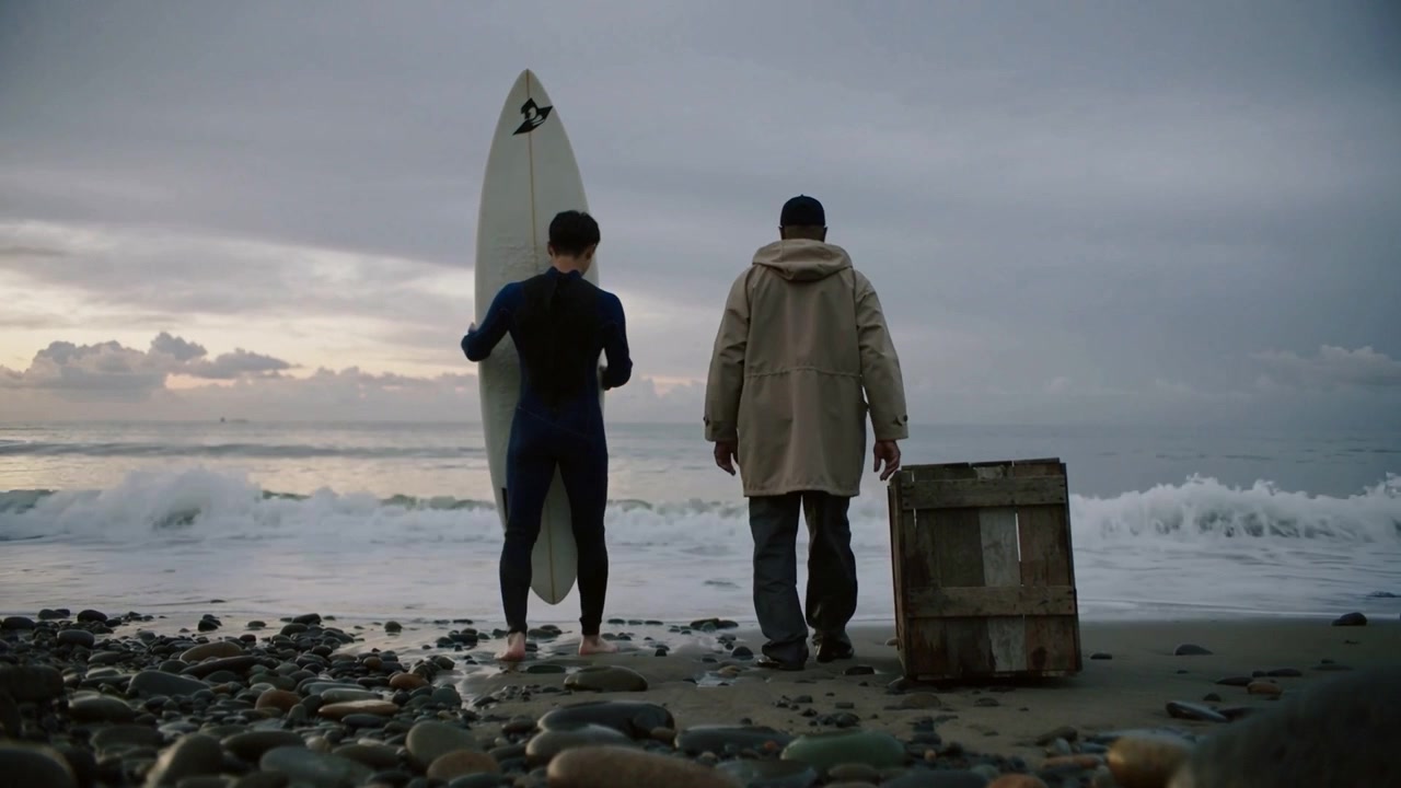 Heading surfer in wetsuit, parka companion into surf on pebbled shore at dawn, carrying board