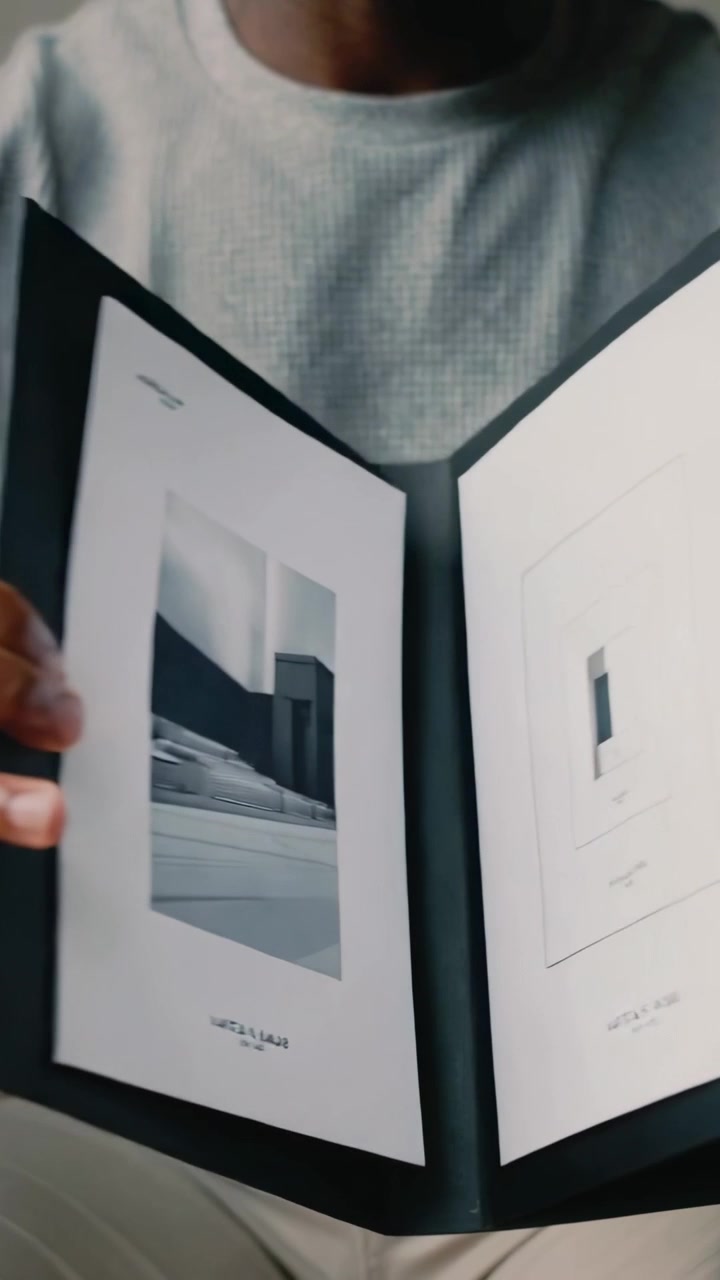 Vertical video: Hands gripping black folio, man in gray shirt opening pages at table showing spread