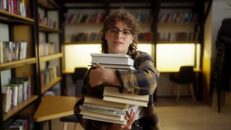 retrato de una estudiante morena con gafas que lleva una pila de libros cerca de los estantes de la biblioteca