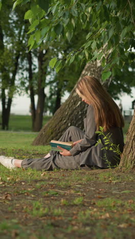 lady in grey clothing sits outdoors under a tree, reading a book with her hand on the page and her leg stretched, the background features greenery and a blur of someone walking in the distance